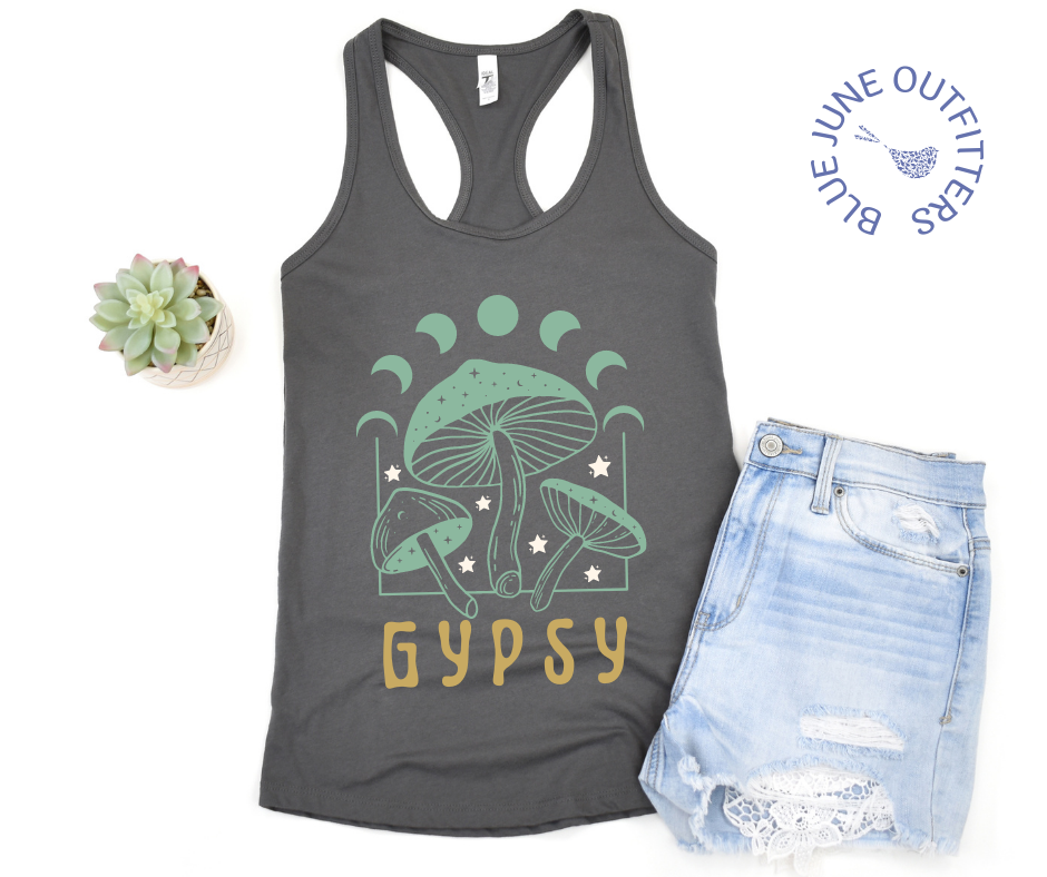 The warm grey tank top lying flat against a white surface, shown here with a pair of distressed denim shorts. The psychedelic artwork is mushrooms, stars and a moon phase with the word GYPSY. The colors of the artwork are mint green, cream and mustard yellow.