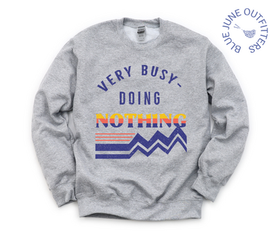 The sport grey sweatshirt on a flat white surface. The artwork is a retro design in dark blue, orange and yellow that reads Very Busy Doing Nothing.