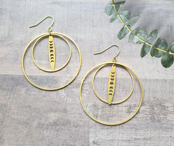 A pair of gold-colored hoop earrings with moon phase charms, displayed on a textured surface.