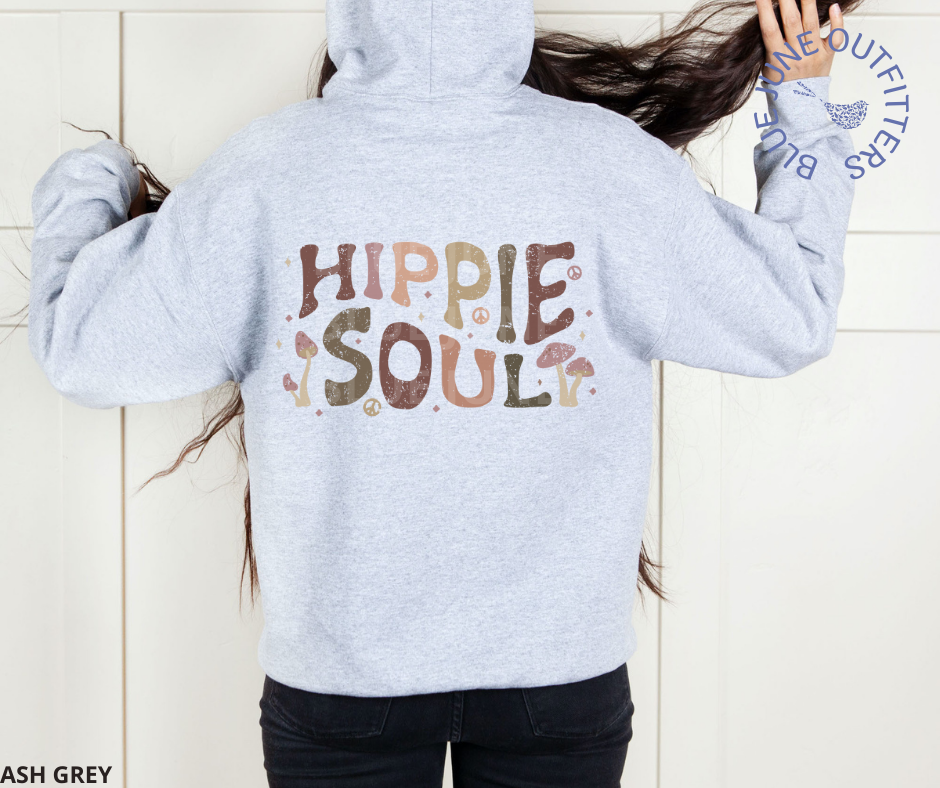 Model wearing the hooded sweatshirt in ash grey. They are facing the wall to show off the back of the hoodie where the artwork is printed. This is from the exclusive Hippie Collection. The text reads Hippie Soul. The font is a retro, wavy text with earthy colors. Small peace signs and wild mushrooms are mixed in around the text.