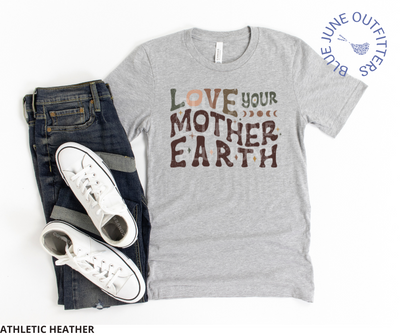 Bella Canvas brand tee in athletic heather. Lying flat with a pair of dark blue jeans and pair of white sneakers. . This shirt is from Blue June Outfitters' exclusive Hippie Collection. It features the phrase love your mother earth. The front is a wavy, retro font in earthy colors. Stars and moon phase are sprinkled within the text.