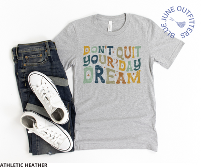 Super soft Bella + Canvas brand tee in athletic heather. Shown here with dark blue jeans. This tee is from Blue June Outfitters' exclusive Hippie Collection. The text reads Don't Quit Your Daydream in hippie wavy letters in earthy blue, yellow and green colors.