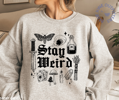 Female modeling the sport grey sweatshirt. Artwork is black. It says stay weird. Surrounding the text are witchy things such as sage, stardust, mushrooms, skull, death potion, dagger, crystals and potions.