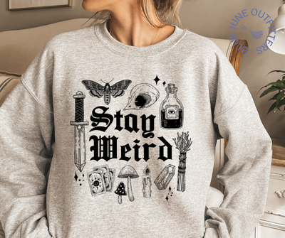 Female modeling the sport grey sweatshirt. Artwork is black. It says stay weird. Surrounding the text are witchy things such as sage, stardust, mushrooms, skull, death potion, dagger, crystals and potions. 