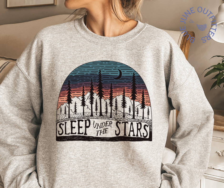 Model wearing an oversized sport grey sweatshirt.  The artwork is an ombre sunset with tall fir trees, mountains and a crescent moon. The text reads SLEEP UNDER THE STARS. 