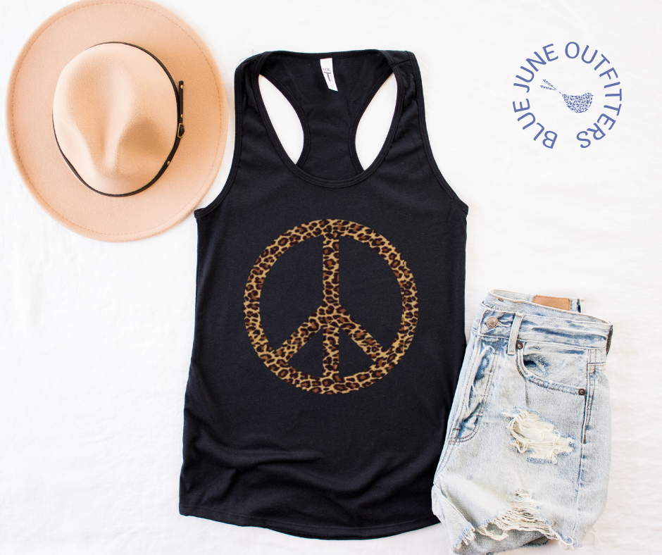 The black women's racerback tank top laid flat on a white surface. Shown here with a pair of jean shorts and trendy hat. The artwork is a leopard pattern peace sign. 
