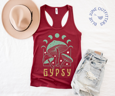 The cardinal red tank top lying flat on a white surface, shown here with a women's summer fedora and distressed denim shorts. The psychedelic artwork is mushrooms, stars and a moon phase with the word GYPSY. The colors of the artwork are mint green, cream and mustard yellow.