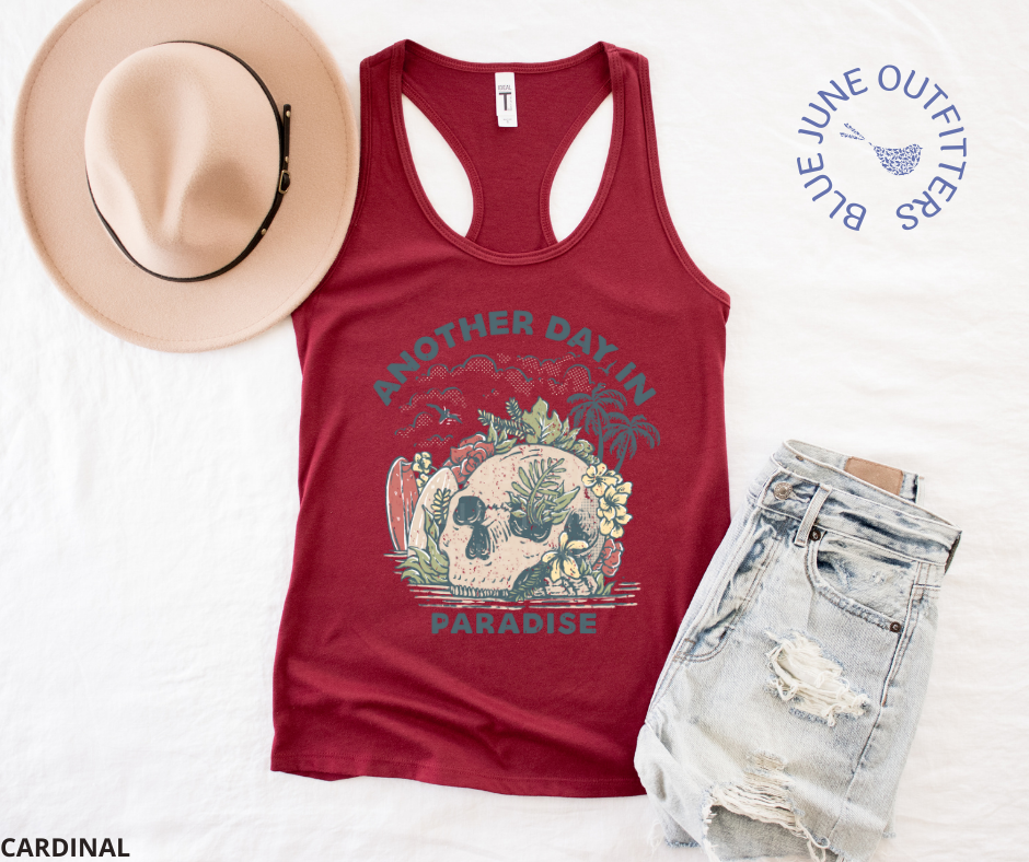 Super soft women's slim fit racerback tank top in cardinal red. Paired here with jean shorts and a trendy hat. This tank is from Blue June Outfitters' exclusive Morbid Nature Collection and features a skull on the beach with palm trees, tropical plants, seagulls and surf boards. The text reads another day in paradise.