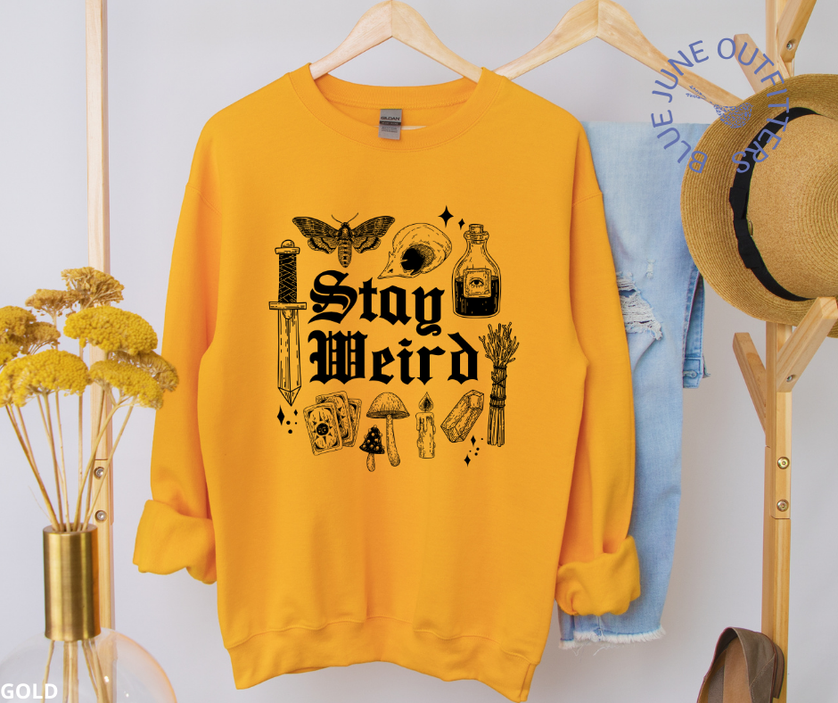 The gold sweatshirt on a clothes hanger next to a vase of yellow flowers and trendy hat.  Artwork is black. It says stay weird. Surrounding the text are witchy things such as sage, stardust, mushrooms, skull, death potion, dagger, crystals and potions.
