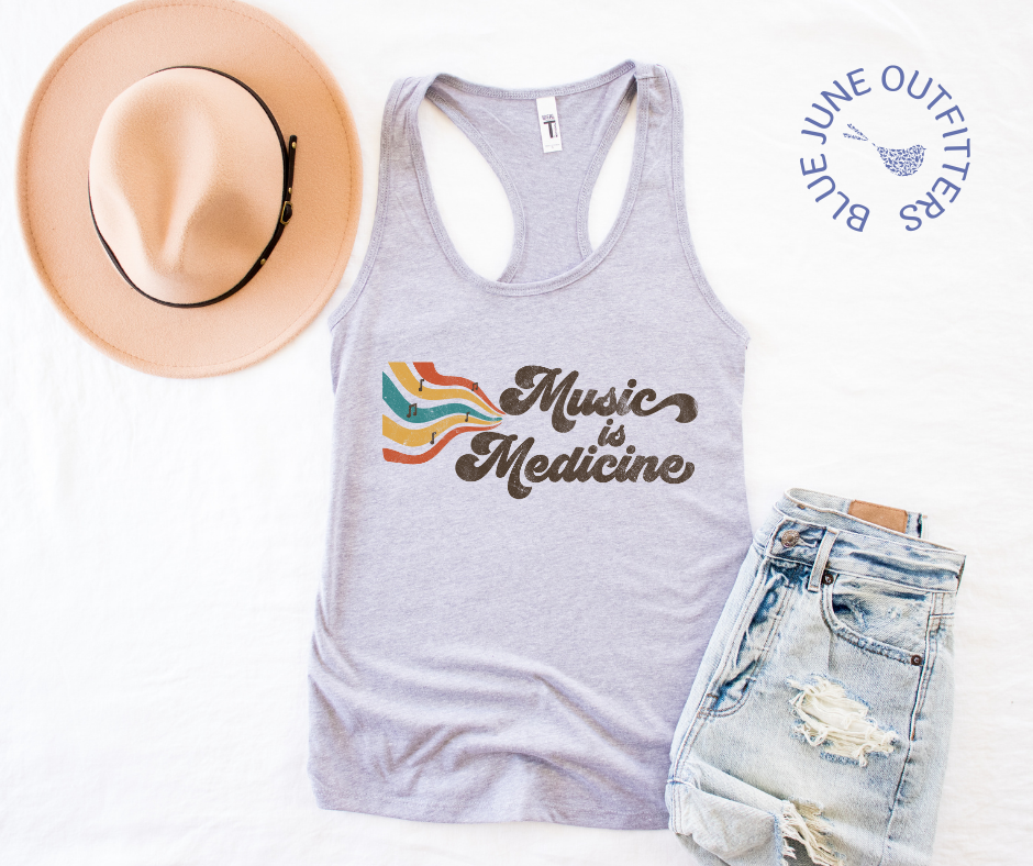 The heather grey racer back tank top lying flat on a white surface.  Pictured here with a trendy hat and distressed jean shorts. The hippie style artwork shows retro music staff and notes with the phrase Music is Medicine. The artwork contains earthy browns, yellows, oranges and turquoise.Music Is Medicine Women's Retro Tank Top