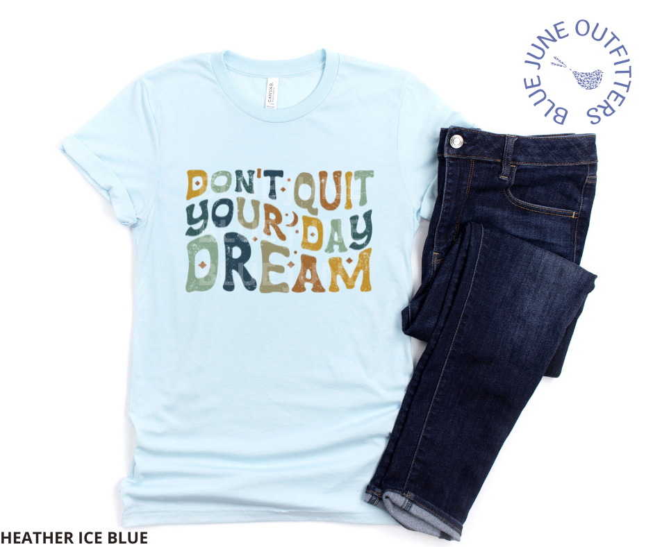 Super soft Bella + Canvas brand tee in heather ice blue. Shown here with dark blue jeans. This tee is from Blue June Outfitters' exclusive Hippie Collection. The text reads Don't Quit Your Daydream in hippie wavy letters in earthy blue, yellow and green colors.
