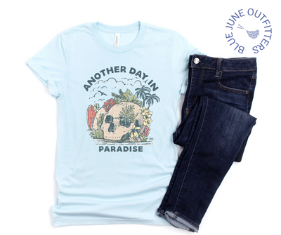 Super soft Bella + Canvas shirt in heather ice blue shown paired with a pair of dark blue jeans. This tee is features a skull on the beach with palm trees, surf boards and seagulls with the text another day in paradise. This is from Blue June Outfitters' exclusive Morbid Nature Collection.