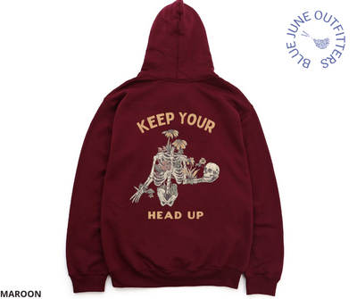 Soft unisex hooded sweatshirt in maroon. This hoodie is from Blue June Outfitters' exclusive Morbid Nature Collection and features a skeleton covered in plants holding his own skull up in his hand. The text reads keep your head up. Artwork and text printed on the back of the hooded sweatshirt.