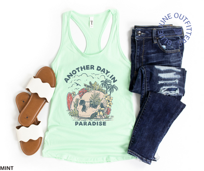 Super soft women's slim fit racerback tank top in mint. Paired here with dark blue jeans and white slide sandals. This tank is from Blue June Outfitters' exclusive Morbid Nature Collection and features a skull on the beach with palm trees, tropical plants, seagulls and surf boards. The text reads another day in paradise.