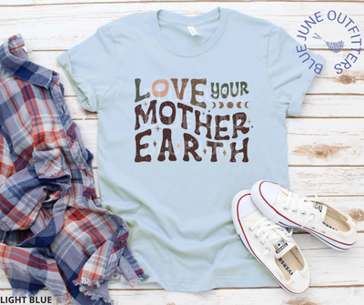 Bella Canvas brand tee in light blue. Lying flat with a flannel shirt and pair of white converse shoes. This shirt is from Blue June Outfitters' exclusive Hippie Collection. It features the phrase love your mother earth. The front is a wavy, retro font in earthy colors. Stars and moon phase are sprinkled within the text.