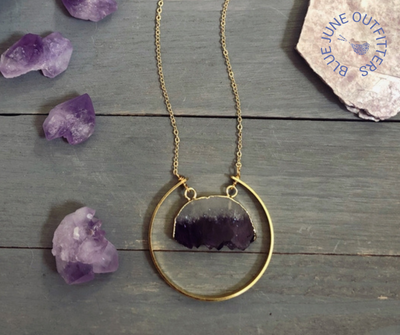 Close view of the raw amethyst necklace. The raw amethyst slice is gold electroplated and surrounded by a raw brass semi-circle. The chain is gold-plated.  Around the necklace are raw amethyst gemstone crystals for a fun accent. 