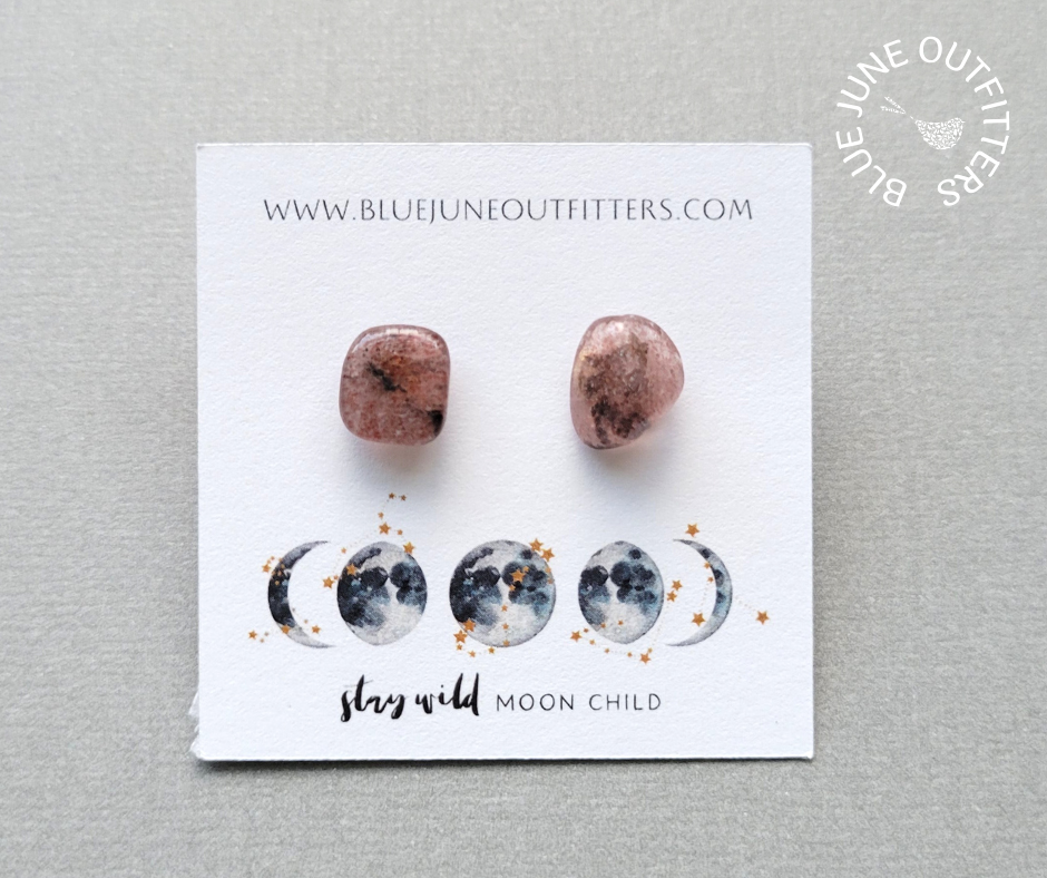 Pair of strawberry quartz stud earrings with beautiful mauve, pink and black coloring. They are organically shaped and on a jewelry card that contains our web address at the top. The bottom of the card is a gold and blue moon phase with the text STAY WILD MOON CHILD on it.