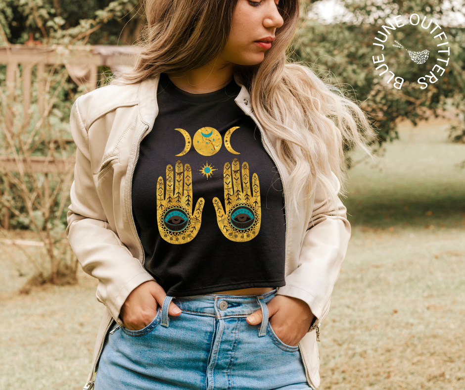 Female wearing the black cropped top. Her midriff shows slightly. The artwork is gold and turquoise. It is two hamsa hands and a moon phase. 
