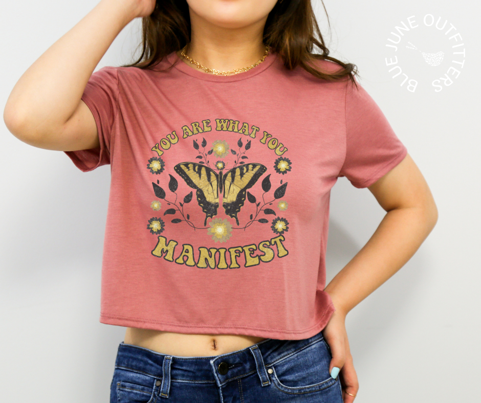 Female wearing the mauve cropped tee. Her midriff shows slightly. The artwork is a vintage style yellow and black butterfly with leaves and flowers. The text reads YOU ARE WHAT YOU MANIFEST. 