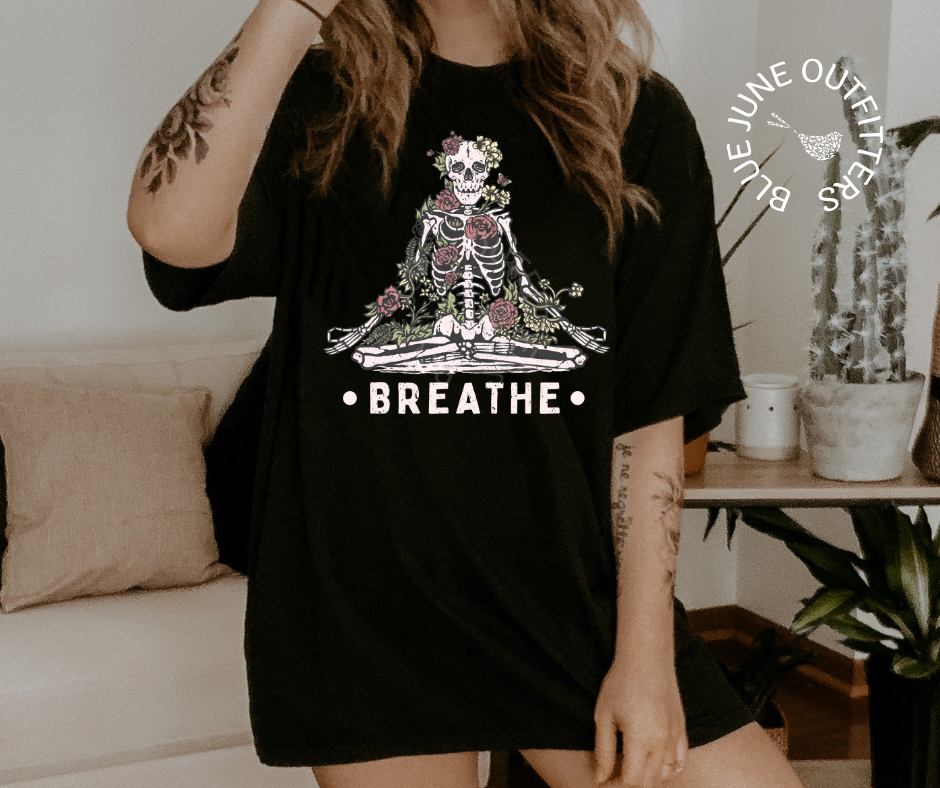 Female with tattoos on the arms wearing the black tee in size XL for an oversized look. A tee from Blue June Outfitters' exclusive Morbid Nature collection with a meditating skeleton surrounded by wildflowers and the phrase breathe printed underneath.