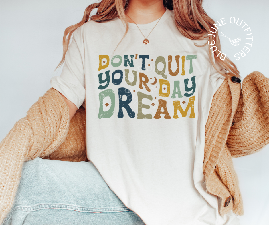 Female modeling the super soft Bella + Canvas brand tee in natural. The text reads Don't Quit Your Day Dream in wavy hippie font. The letters are in earthy yellow, green, blue and orange tones.