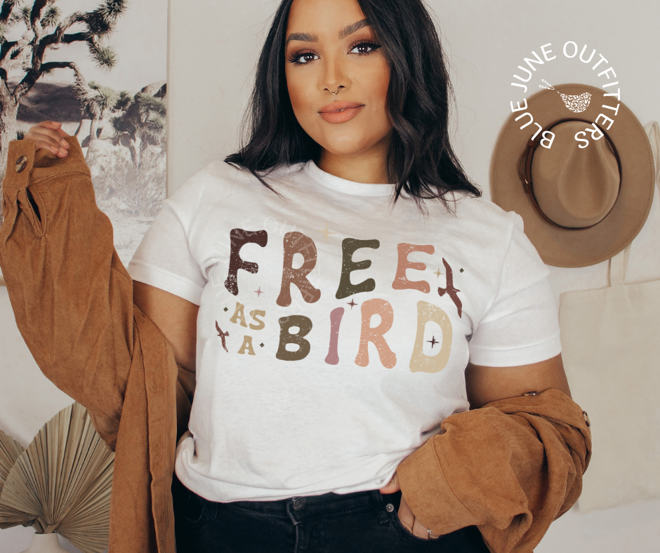 Ethnic model wearing the white t-shirt with an oversized suede jacket. This shirt is from Blue June Outfitters exclusive Hippie Collection The shirt reads Free As A Bird. It is a trippy retro font in earthy colors. There are small birds in flight and stars around the text.