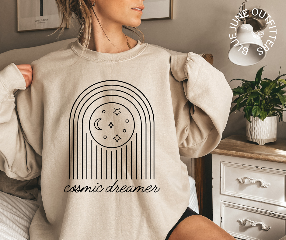 Female modeling an oversized sweatshirt in sand color. The image on the sweatshirt is of stars and moons. In cursive, the text reads cosmic dreamer.