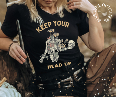Female modeling the black tee with a witchy celestial skirt. This shirt from Blue June Outfitters' exclusive Morbid Nature Collection features a skeleton covered in plants holding his skull up in his hands with the phrase keep your head up. This is the perfect tee for those who share