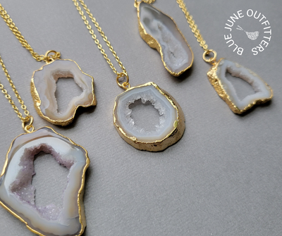 View of five different electroplated agate druzy slice pendants. They are various size and shapes. All are a creamy lavender, bream and brown color. They are attached to a gold chain. 