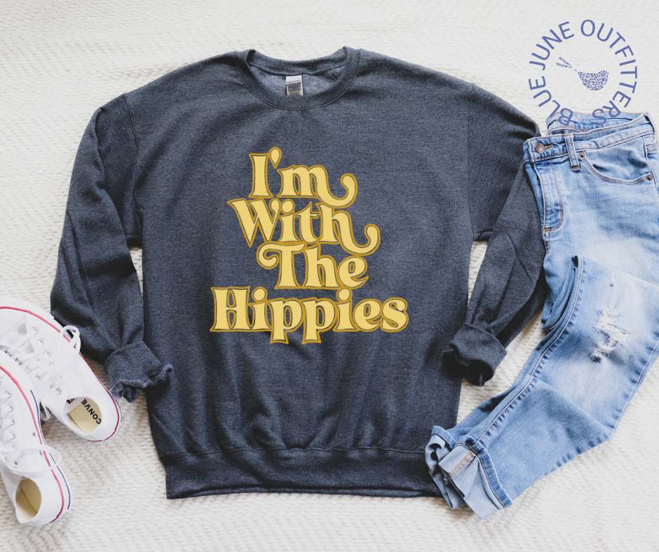 The dark heather crewneck sweatshirt laying flat on a white surface, shown here with a pair of faded and distressed jeans and a pair of white converse sneakers. The text is a mustard yellow, retro wavy font that reads I'M WITH THE HIPPIES.