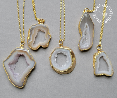 View of five different electroplated agate druzy slice pendants. They are various size and shapes. All are a creamy lavender, bream and brown color. They are attached to a gold chain.