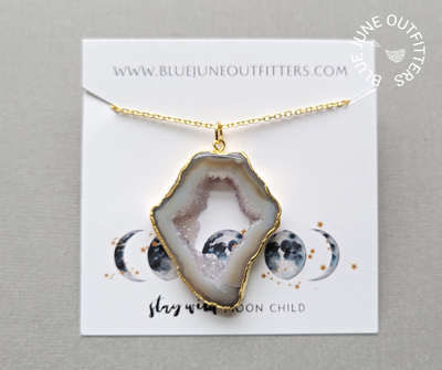 A photo of the agate slice druzy necklace presented on a jewelry card which is how it comes packaged. The card reads www.bluejuneoutfitters.com at the top and at the bottom are gold and blue moon phases with the phrase, STAY WILD MOON CHILD. 
