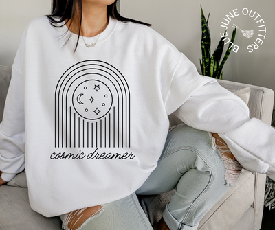 Female modeling an oversized sweatshirt in white. The image on the sweatshirt is of stars and moons. In cursive, the text reads cosmic dreamer. 