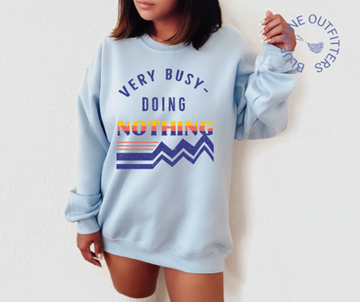 Female modeling an oversized sweatshirt in light blue. The artwork is a retro design in dark blue, orange and yellow that reads Very Busy Doing Nothing. 