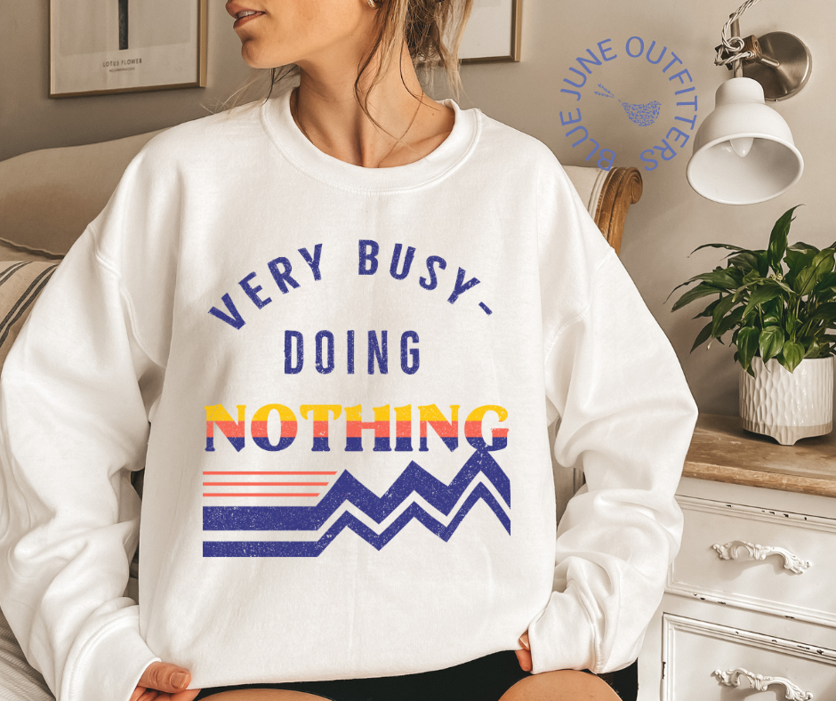 Female modeling an oversized sweatshirt in white. The artwork is a retro design in dark blue, orange and yellow that reads Very Busy Doing Nothing.