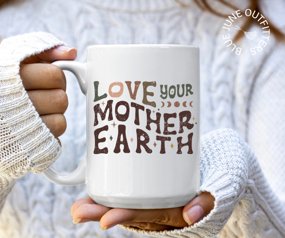 15 oz white ceramic mug with c-handle and rounded lip. Being held here by a model wearing a cozy white sweater. The artwork says love your mother earth. The letters are hippie style wavy letters in dark earth tones of greens, browns and peach. There are stars and moons accents. 
