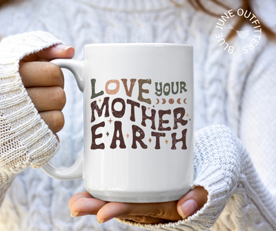 15 oz white ceramic mug with c-handle and rounded lip. Being held here by a model wearing a cozy white sweater. The artwork says love your mother earth. The letters are hippie style wavy letters in dark earth tones of greens, browns and peach. There are stars and moons accents. 