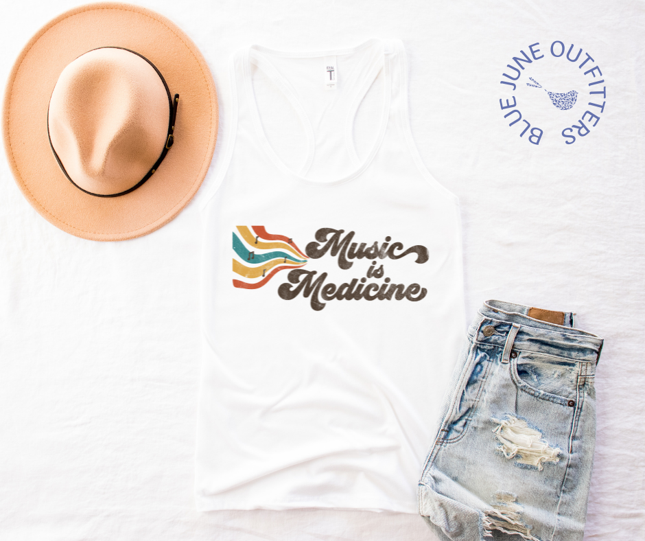 The white racerback tank top lying flat on a white surface. Pictured here with a trendy hat and distressed jean shorts. The hippie style artwork shows retro music staff and notes with the phrase Music is Medicine. The artwork contains earthy browns, yellows, oranges and turquoise.Music Is Medicine Women's Retro Tank Top