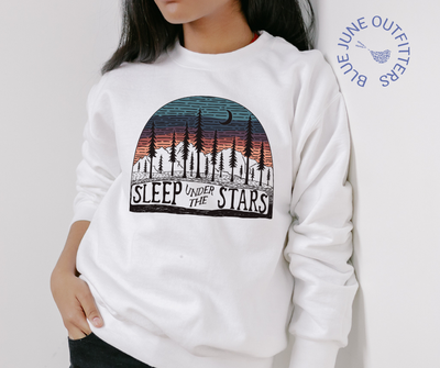 Model wearing the white sweatshirt. The artwork is an ombre sunset with tall fir trees, mountains and a crescent moon. The text reads SLEEP UNDER THE STARS.