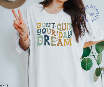 Female modeling the super soft Bella + Canvas brand tee in white. The text reads Don't Quit Your Day Dream in wavy hippie font. The letters are in earthy yellow, green, blue and orange tones. 