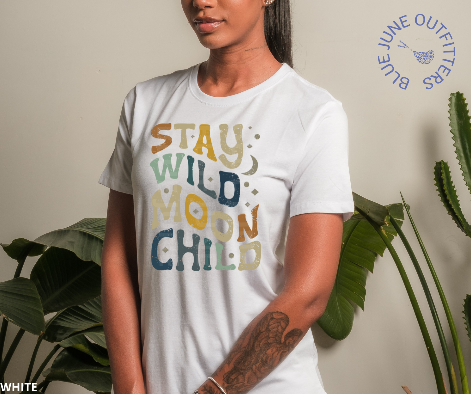 Woman stands in front of a light grey wall with house plants on each side of her. She is looking to the left and holds her hands clasped in front of her. Unisex Bella + Canvas tee in ash grey. This shirt is from our exclusive Hippie Collection and features wavy, hippy letters that rea stay wild moon child. There are also small stars and a crescent moon intermixed with the lettering. The colors of the text are a mix of dark blue, earthy green, mustard yellow and rust.