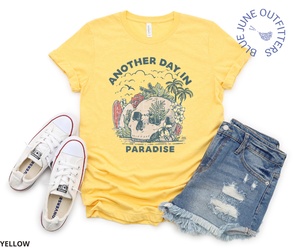 Super soft Bella + Canvas shirt in yellow, shown here with jean shorts and white converse tennis shoes. This tee is features a skull on the beach with palm trees, surf boards and seagulls with the text another day in paradise. This is from Blue June Outfitters' exclusive Morbid Nature Collection.