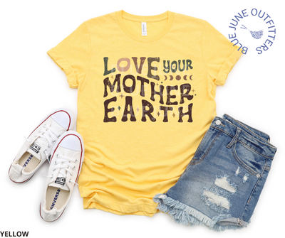Bella Canvas brand tee in yellow. Lying flat here with a pair of distressed denim shorts and a pair of white converse sneakers. This shirt is from Blue June Outfitters' exclusive Hippie Collection. It features the phrase love your mother earth. The front is a wavy, retro font in earthy colors. Stars and moon phase are sprinkled within the text.