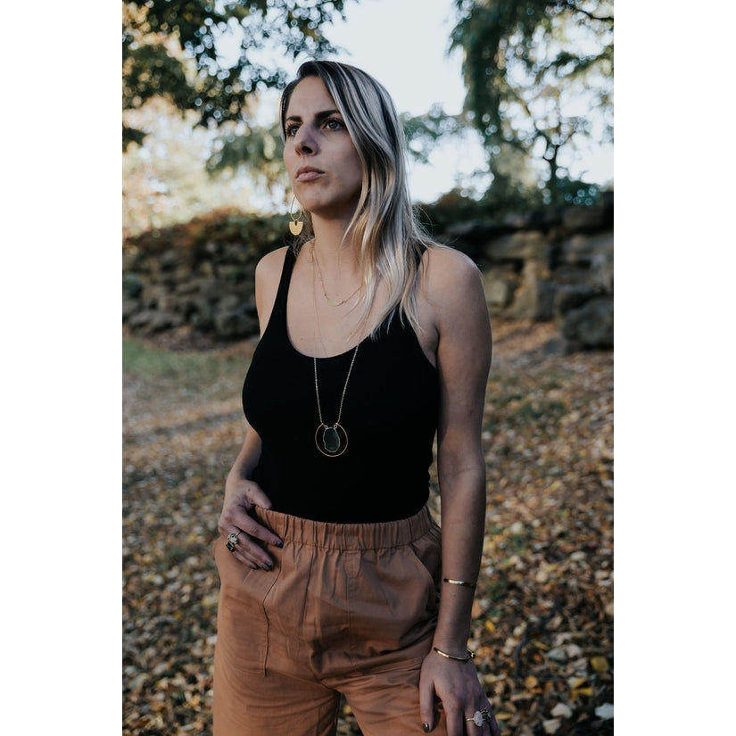 Female modeling the necklace in nature.  She wearing a black tank top with rust orange slacks. The necklace pairs beautifully and the pendant sits just under the bust. 