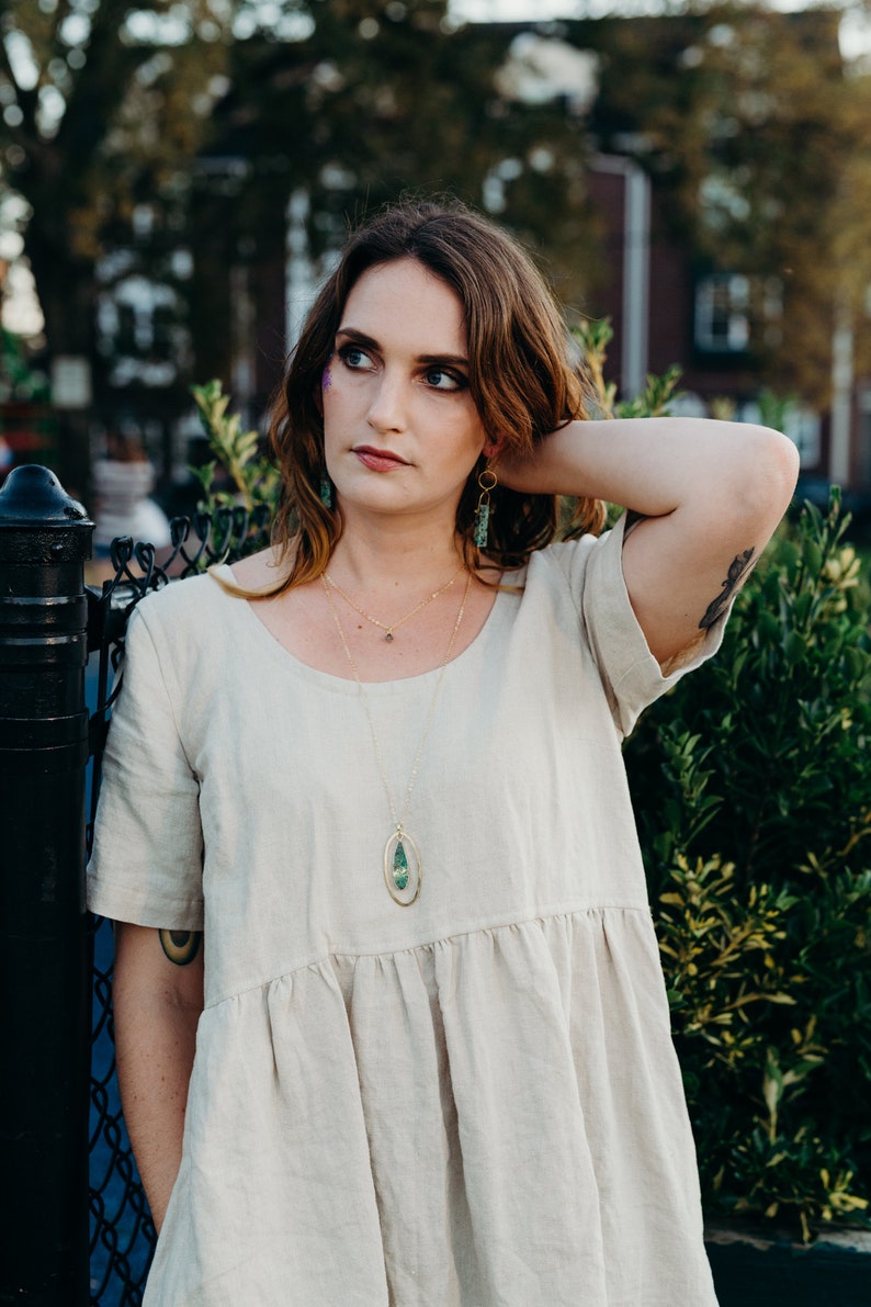 Female modeling the necklace. She's standing outside next to plants, wearing a tan frock.  The pendant falls just below the bust line. 