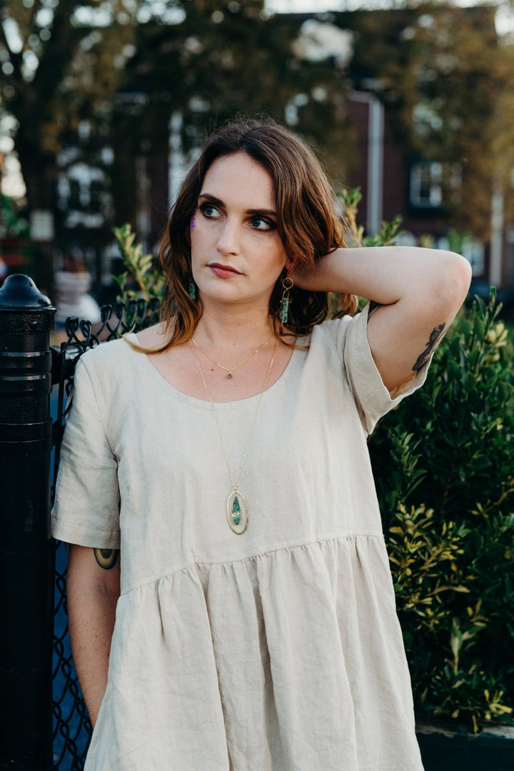 Female modeling the necklace. She's standing outside next to plants, wearing a tan frock.  The pendant falls just below the bust line. 