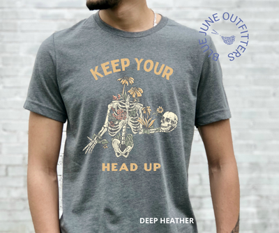 Super soft Bella + Canvas tee in deep heather grey. This shirt from Blue June Outfitters' exclusive Morbid Nature Collection features a skeleton covered in plants holding his skull up in his hands with the phrase keep your head up. This is the perfect tee for those who share