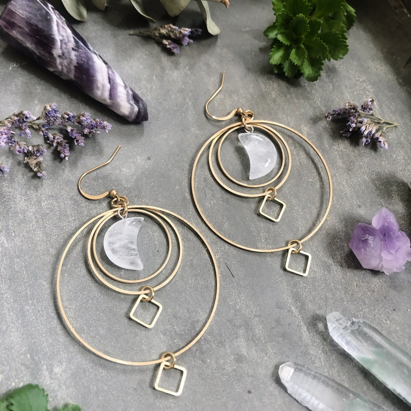 Pair of gypsy style brass dangle earrings, each with clear quartz crystals shaped like crescent moons. There are three brass circles of different diameters and two small diamond shaped brass dangles. The brass ear hooks are hypo allergenic.