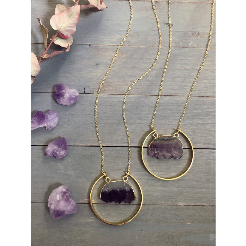 Overhead view of two raw amethyst necklace. The raw amethyst slice is gold electroplated and surrounded by a raw brass semi-circle. The chain is gold-plated. Around the necklace are raw amethyst gemstone crystals for a fun accent.