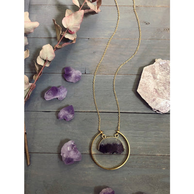 Overhead view of the raw amethyst necklace. The raw amethyst slice is gold electroplated and surrounded by a raw brass semi-circle. The chain is gold-plated. Around the necklace are raw amethyst gemstone crystals for a fun accent.